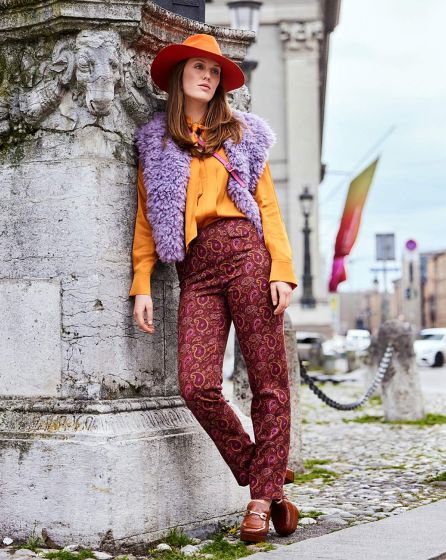 Une femme est appuyée contre une large colonne carrée en pierre d'allure ancienne, au bord d'une rue. Elle porte un pantalon en velours bordeaux imprimé d'un motif cachemire, un chemisier orange, un gilet sans manches en moumoute mauve, et un chapeau orange à larges bords. Elle regarde au loin avec un air pensif, se demandant sans doute ce qu'elle a fait pour mériter ça.